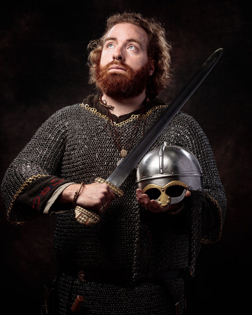 A bearded man in chainmail armour holds a sword and helmet against a dark background, dressed in medieval warrior attire.