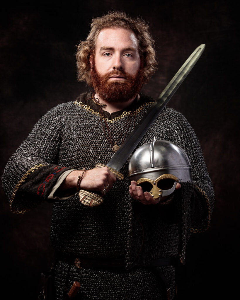 A bearded man in medieval chainmail and gold jewellery holds a sword and helmet against a dark background.