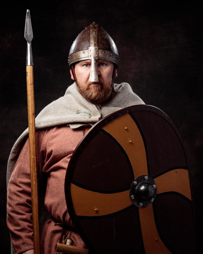A bearded man wearing Viking-era armor, including a metal helmet and shield, holds a spear against a dark background.