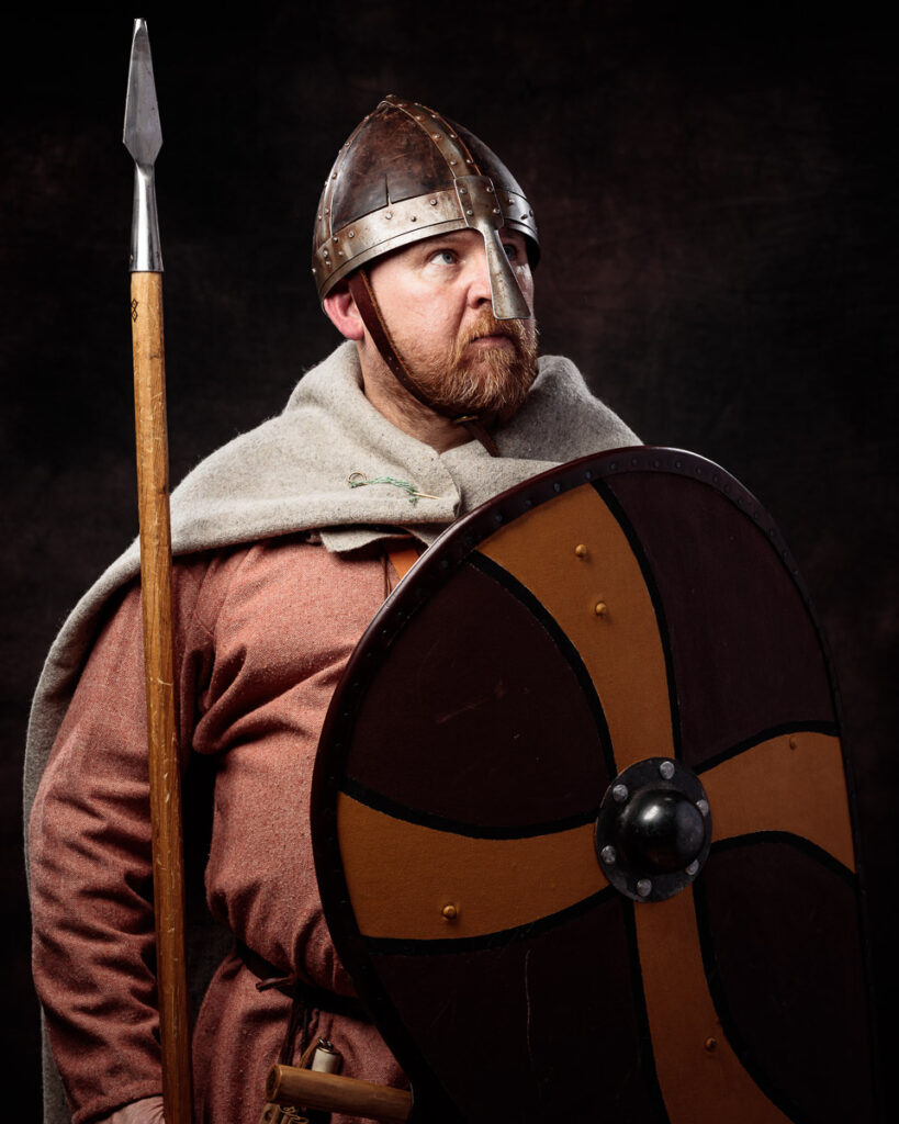 A bearded man in Viking-era clothing holds a spear and shield against a dark background.