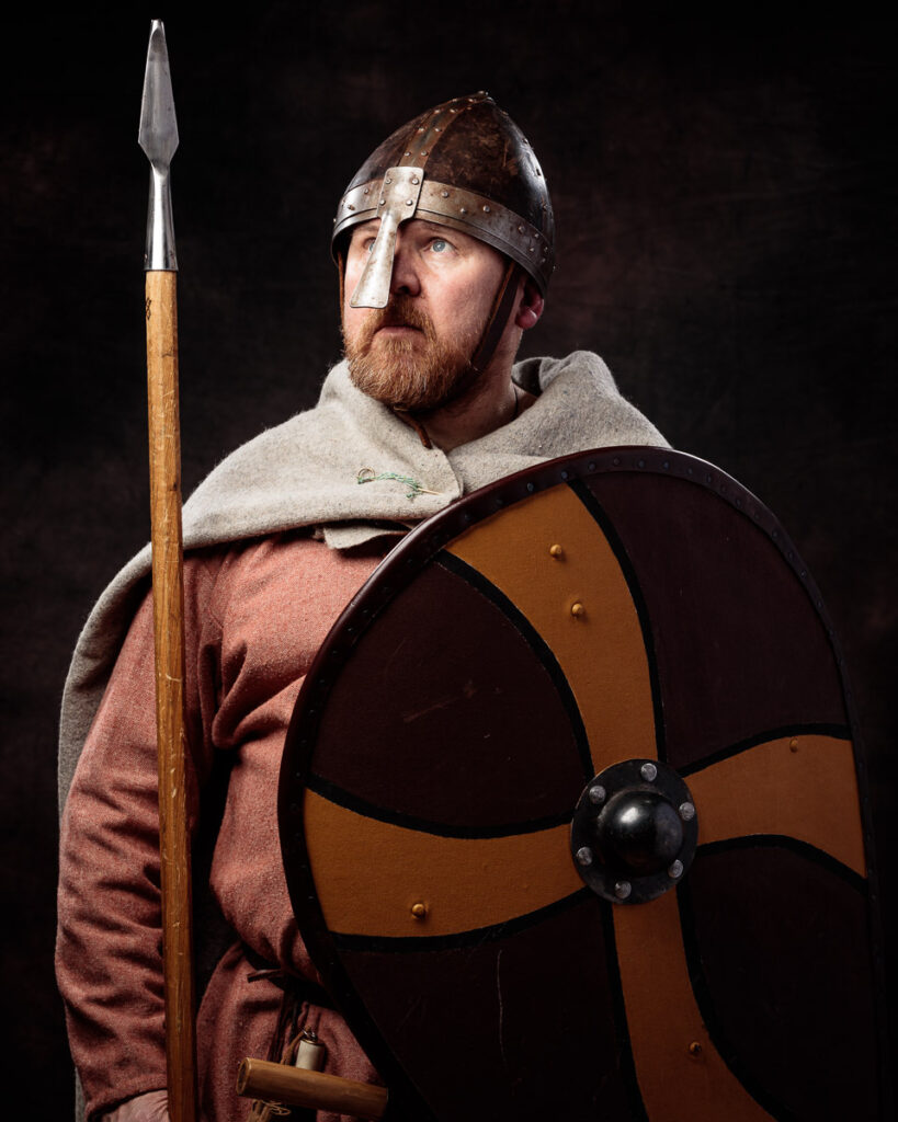 A bearded man dressed as a Viking warrior holds a spear and shield against a dark background.