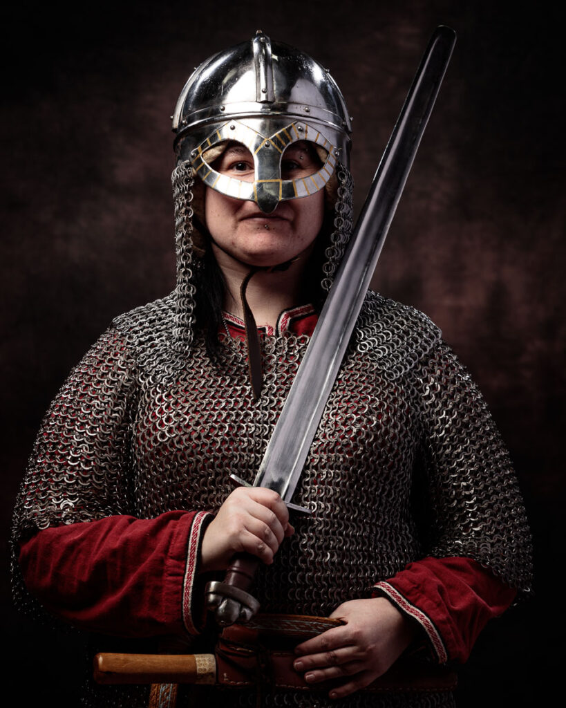 A person in medieval warrior costume wearing a polished metal helmet with chainmail, holding a sword, against a dark background.