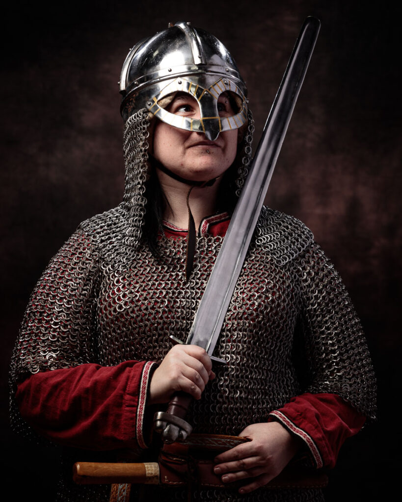 A person in Viking warrior costume wearing a polished metal helmet with chainmail, red tunic and holding a sword against a dark background.