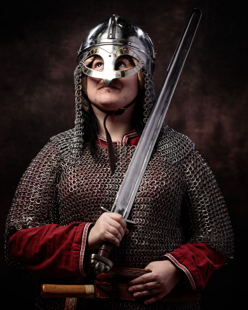 A person in Viking warrior costume wearing a metal helmet with chainmail braids, holding an axe against a dark background.