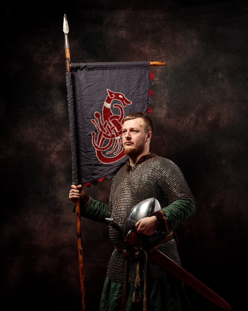 A bearded man in medieval chainmail and green tunic holds a banner with a red dragon emblem and a spear against a dark backdrop.