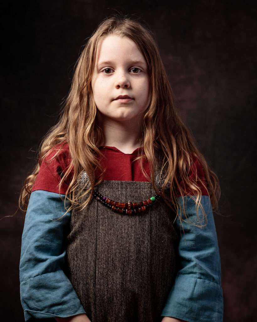 A young girl with long reddish-brown hair wearing a blue and red colour-block top with a beaded necklace, against a dark background.