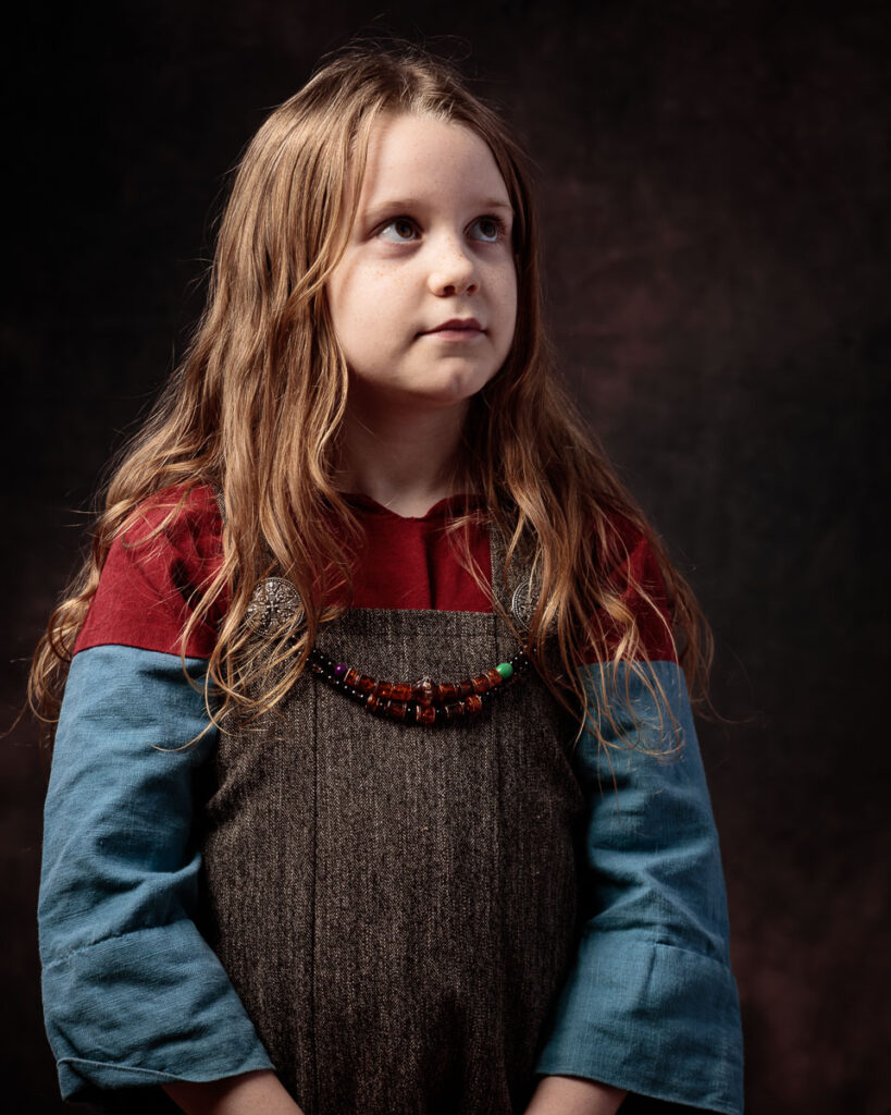 A young girl with long reddish-brown hair wears a medieval-style tunic with blue sleeves and a beaded necklace, gazing to the side against a dark background.