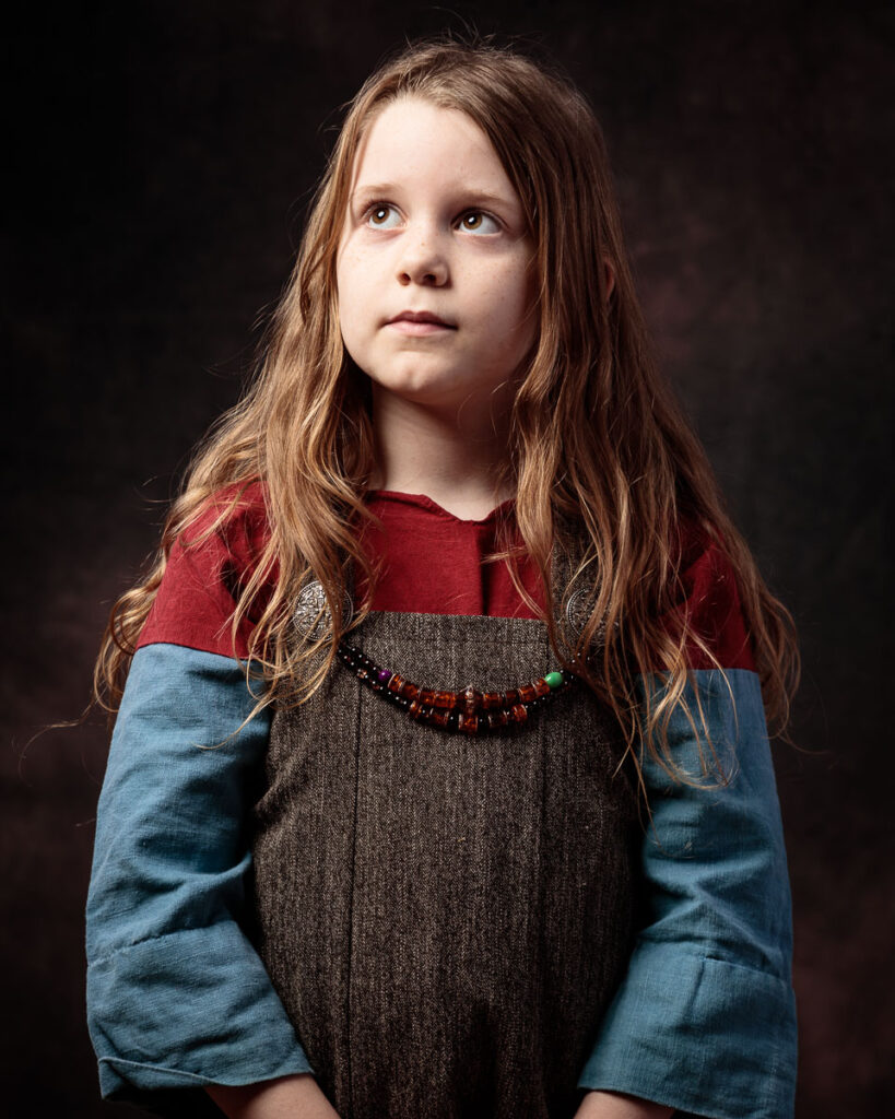 A young girl with long reddish-brown hair wearing a grey tunic with red and blue sleeves and a beaded necklace, looking upward against a dark background.