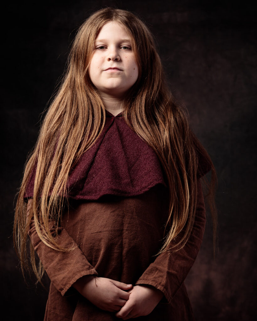 A young girl with long reddish-brown hair wearing a burgundy and brown layered top, photographed against a dark background.