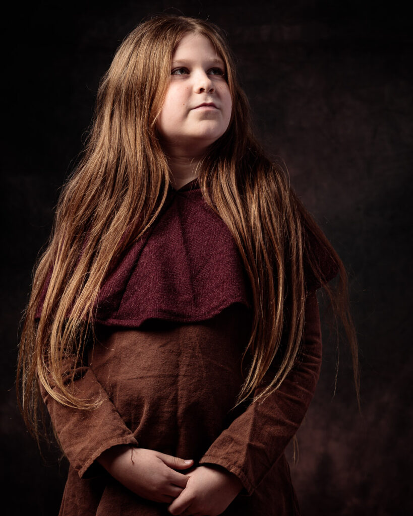 A young girl with long brown hair wearing a burgundy and brown layered top, posed against a dark background.