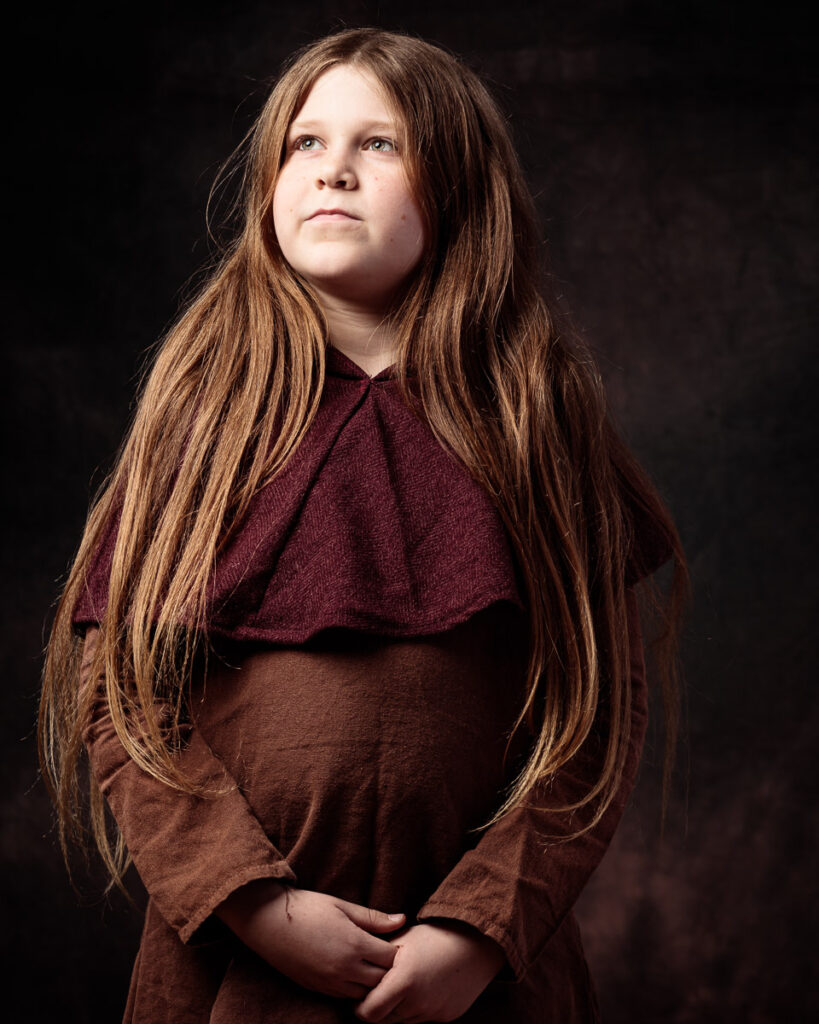 A young girl with long brown hair wearing a burgundy and brown layered top, looking upward against a dark background.