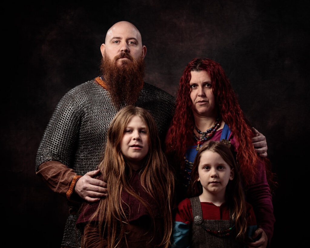 A family of four posed against a dark background. A bearded man wears chainmail and brown tunic. A woman with long red hair wears medieval-style clothing. Two girls with brown hair stand in front wearing burgundy and grey garments.