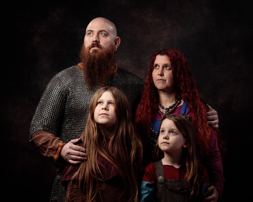A family of four posed against a dark background. An adult man with a long beard wears chainmail. Two young girls and an adult woman with long red hair stand beside him in medieval-style clothing.