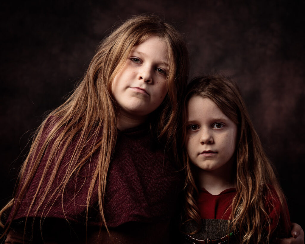 Two young girls with long reddish-brown hair pose together against a dark background, wearing burgundy clothing and looking at the camera.