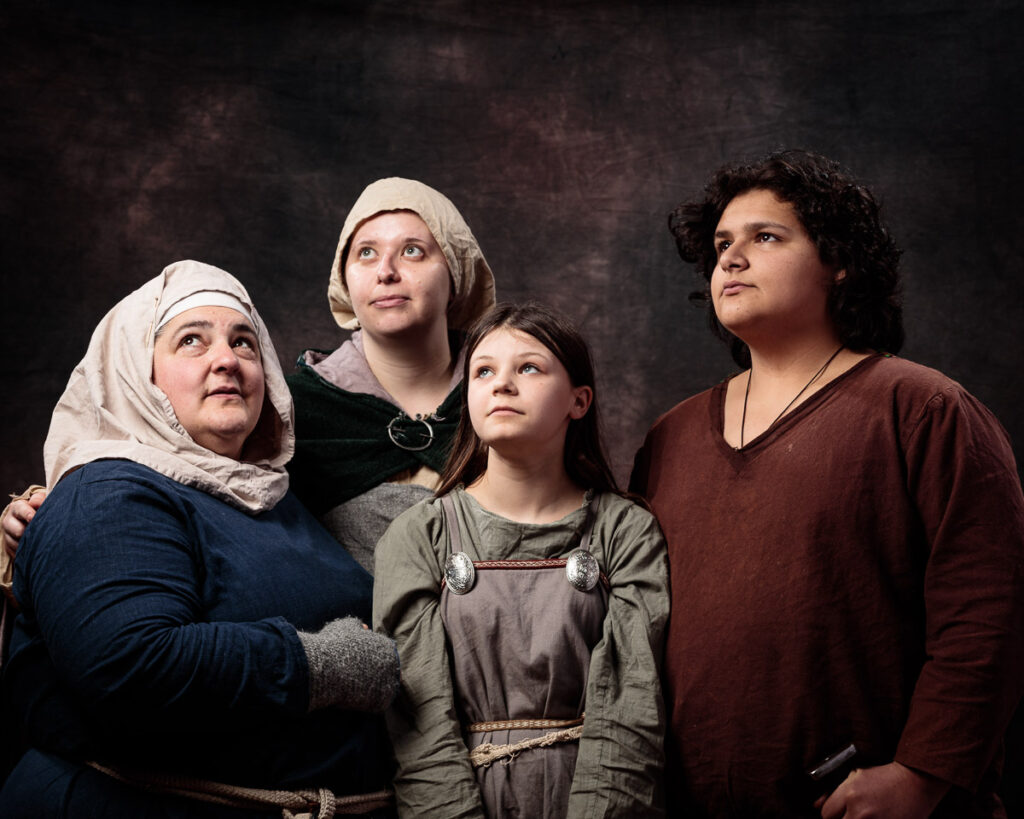 Four people in medieval-style clothing gaze upward against a dark textured backdrop. They wear linen headdresses and earth-toned garments with decorative elements.