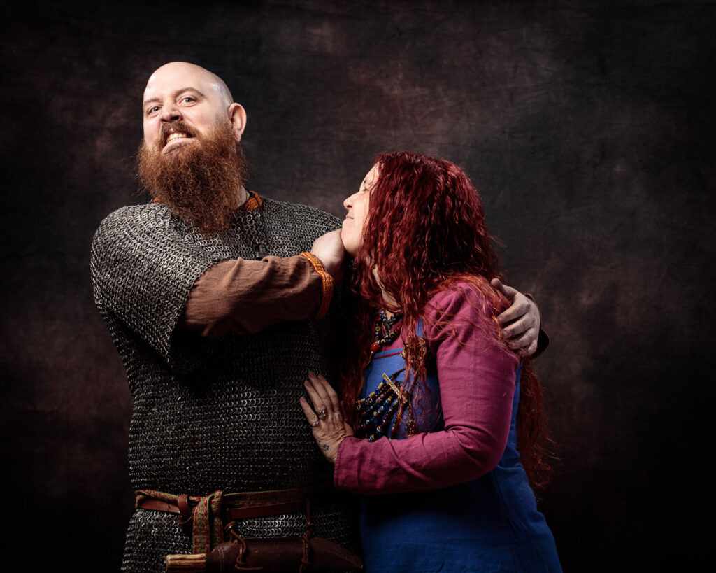 Two people in medieval costume embrace against a dark background. The man wears chainmail and a long beard; the woman has long red hair and a purple tunic.