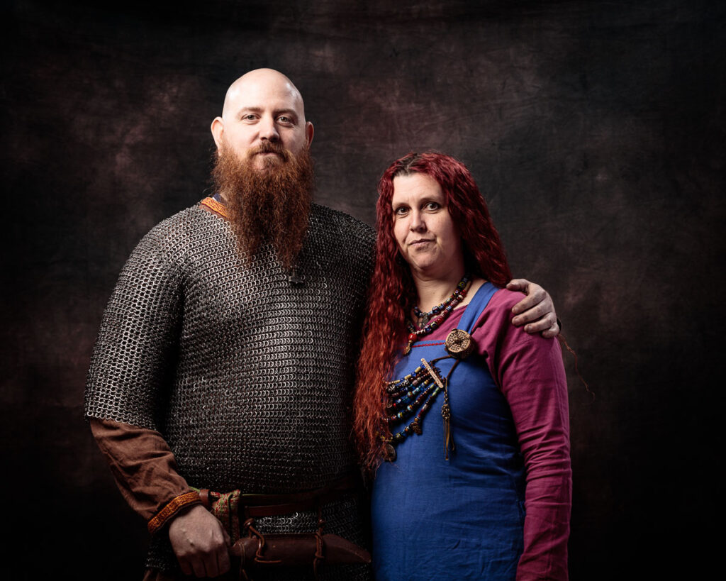 Two people in Viking-era clothing pose together against a dark textured background. The man wears chainmail and brown tunic; the woman wears a blue dress with purple overdress and jewellery.