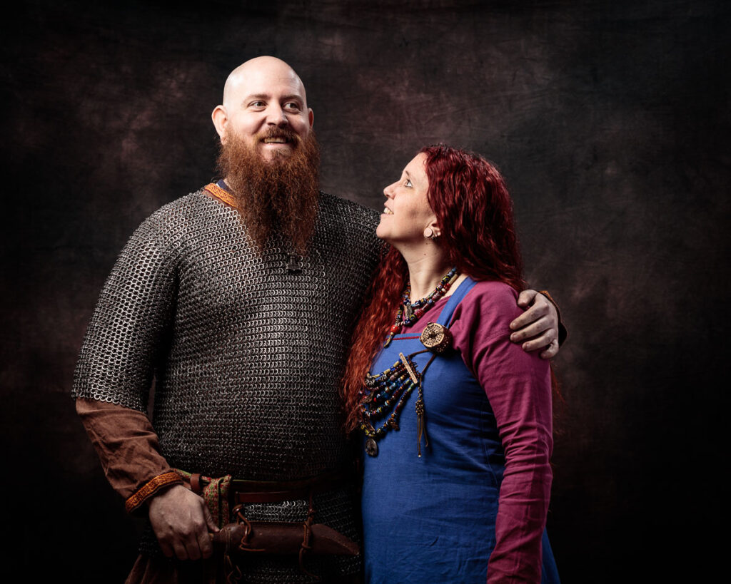 Two people in Viking-era clothing pose together against a dark textured background. The man wears chainmail and brown tunic; the woman wears a blue dress with burgundy overdress and has long red hair.