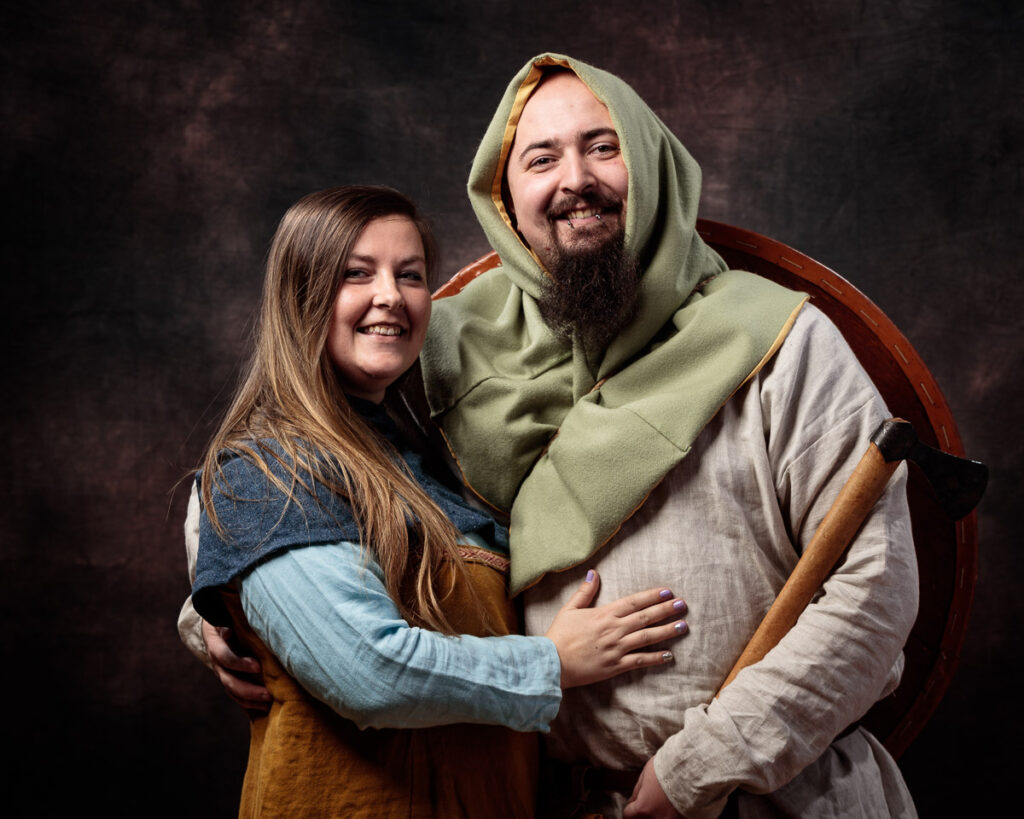 Two people in medieval-style clothing embrace and smile at the camera against a dark textured backdrop.