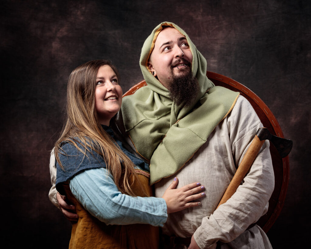 Two people in medieval-style clothing embrace against a dark backdrop, both smiling and looking upward.