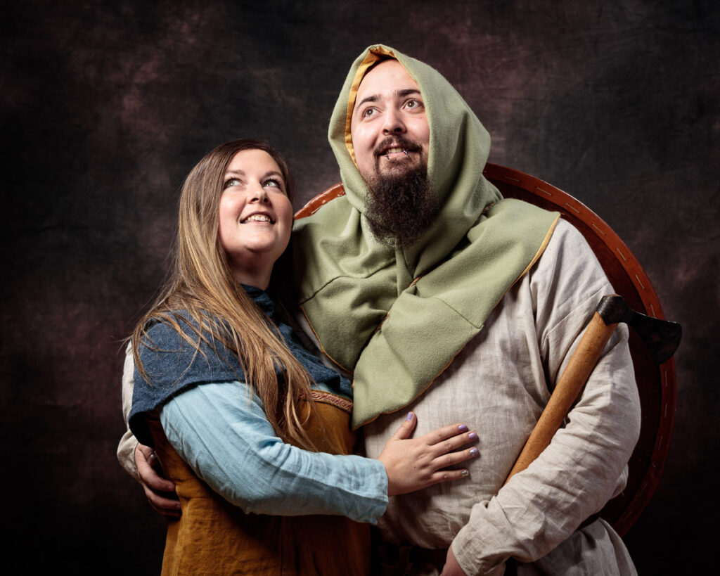 Two people in medieval-style clothing embrace and look upward against a dark textured background.