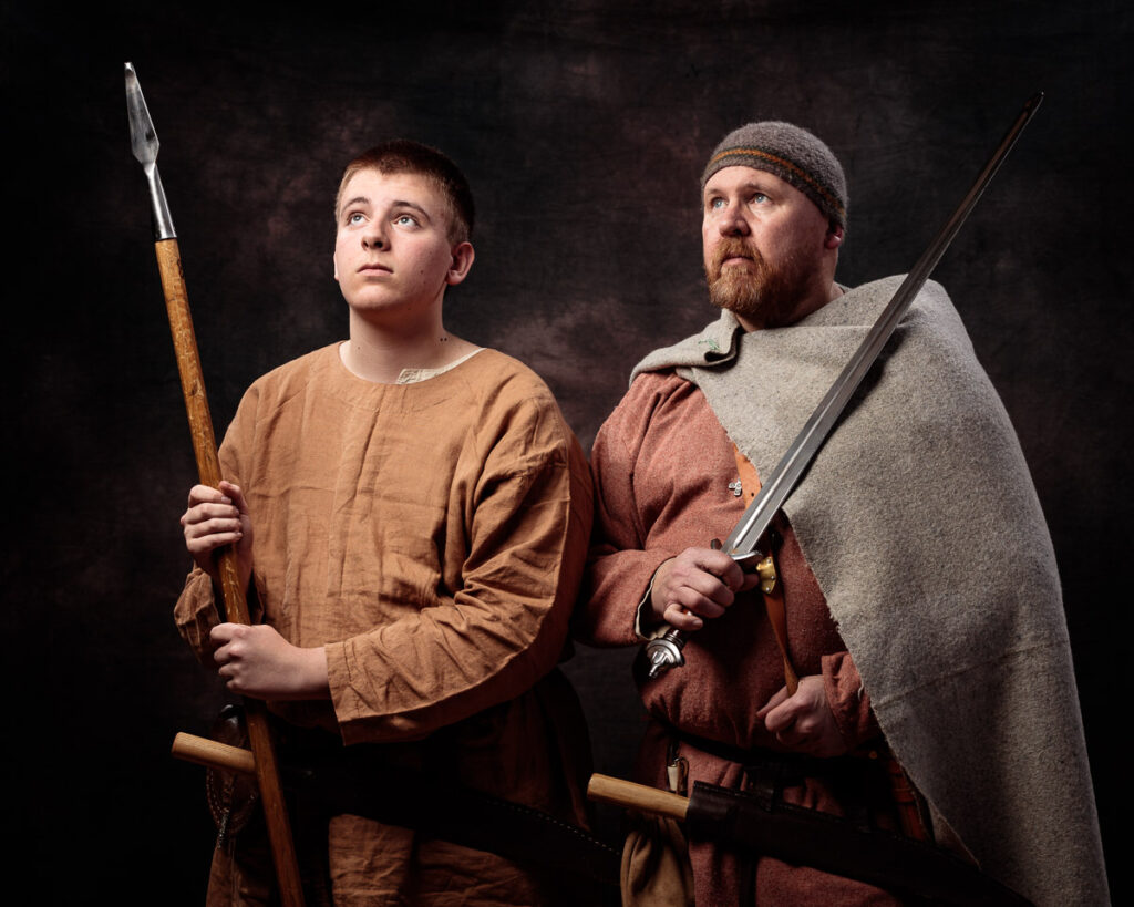 Two men in Viking-era clothing hold weapons against a dark background. The younger man holds a spear, the older wears a beanie and holds a sword.