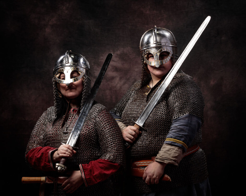 Two people in Viking warrior costumes with metal helmets and chainmail armor hold large swords against a dark background.