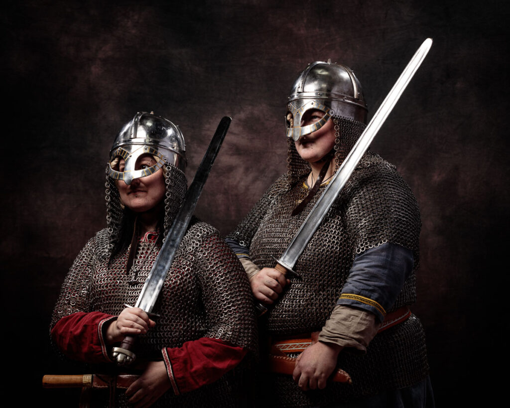 Two armoured warriors in chainmail and metal helmets hold swords against a dark textured background.