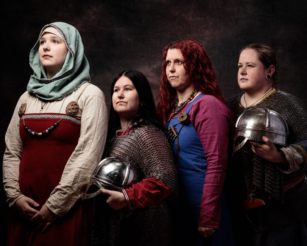 Four women in Viking-era clothing and armour stand against a dark background, holding helmets, gazing upward.