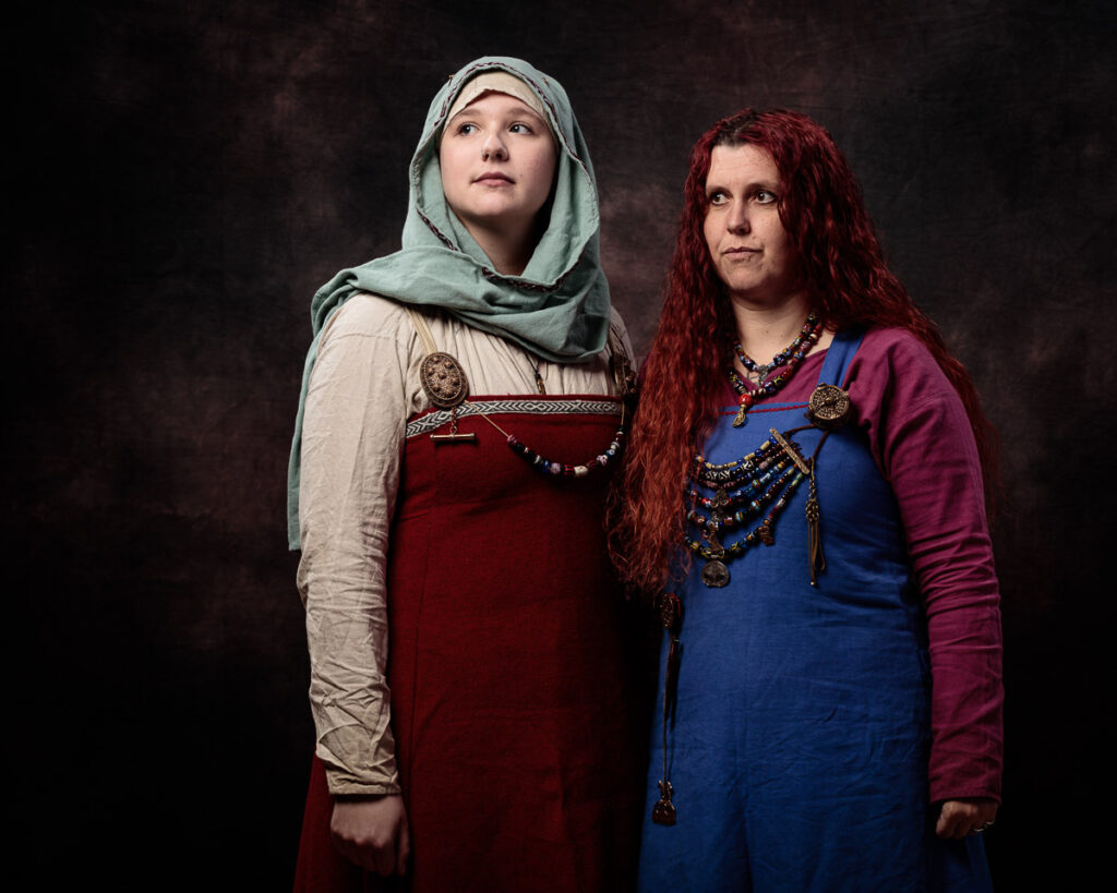 Two women in medieval Slavic dress stand against a dark background. The woman on the left wears a red dress with a grey hood; the woman on the right has long red hair and wears a blue dress with purple sleeves and ornate beaded necklaces.