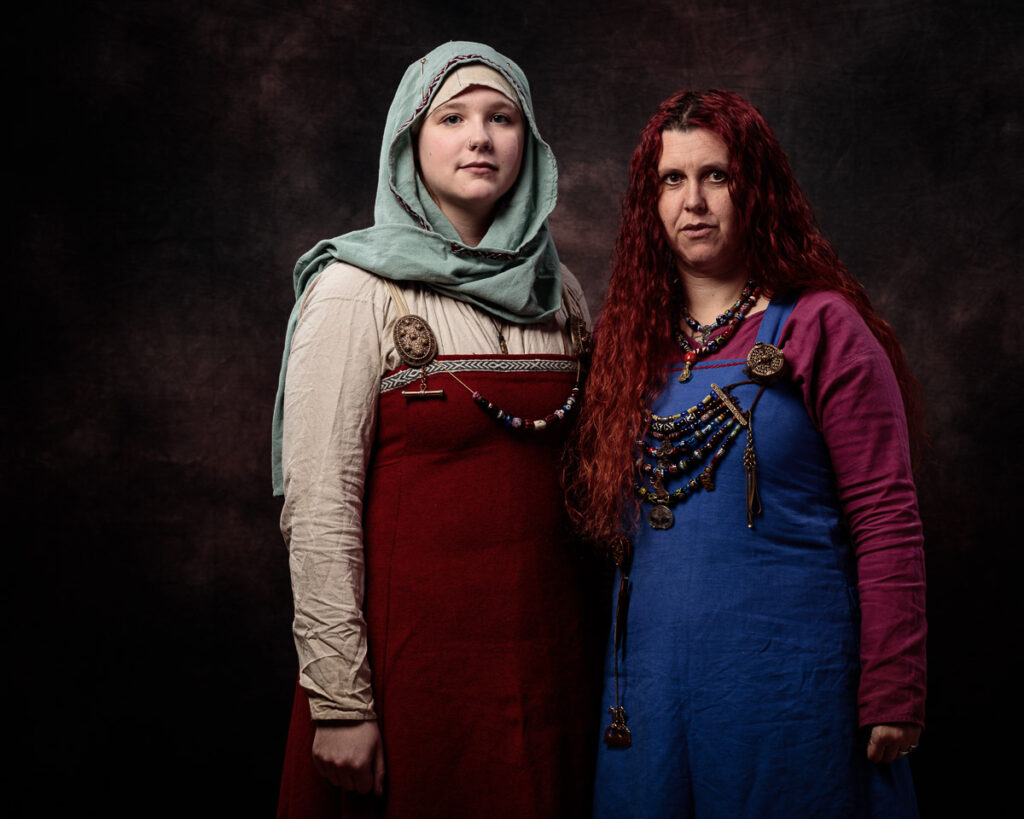 Two women in medieval Slavic costume stand against a dark background. The woman on the left wears a red dress with a grey headscarf. The woman on the right wears a blue dress with a burgundy overdress and has long red hair.