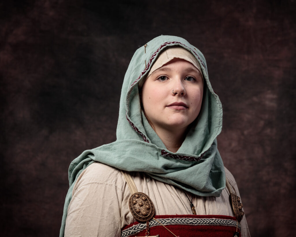 A young woman wearing historical Slavic clothing with a light green linen headscarf and embroidered tunic, gazing upward against a dark brown background.