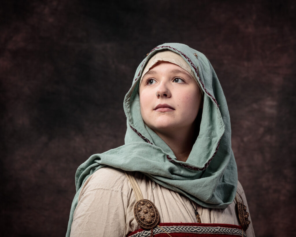 A young woman wearing a grey-green head covering and cream tunic with decorative metal brooch, gazing upward against a dark background.