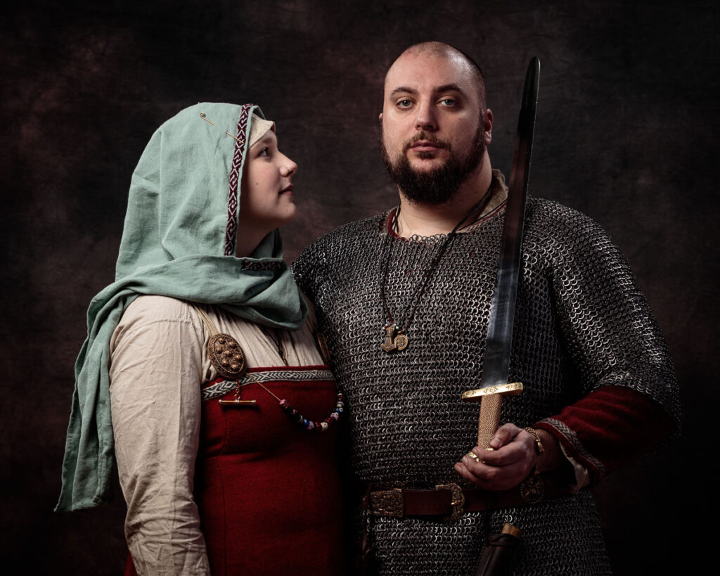 Two people in medieval clothing pose against a dark background. The man wears chainmail and holds a sword; the woman wears a headscarf and red tunic.