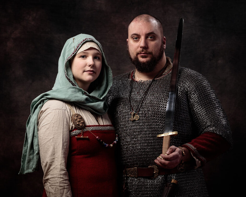 Two people in Viking-era clothing pose against a dark brown background. The man wears chainmail and holds a sword; the woman wears a headscarf and red tunic.