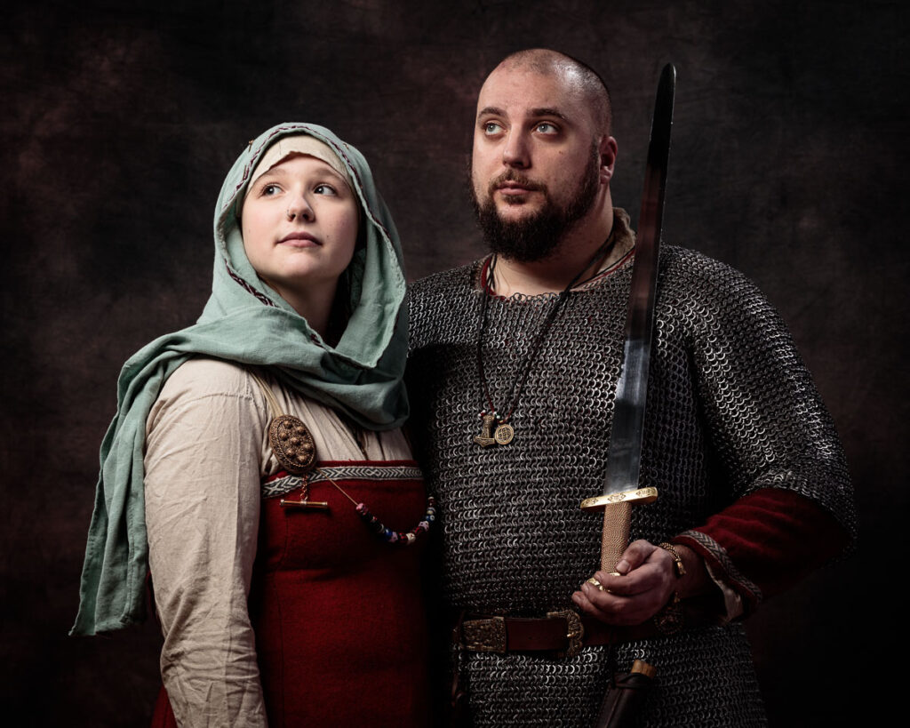 Two people in Viking-era costume pose together against a dark brown background. The man wears chainmail and holds a sword; the woman wears a red dress and green head covering.