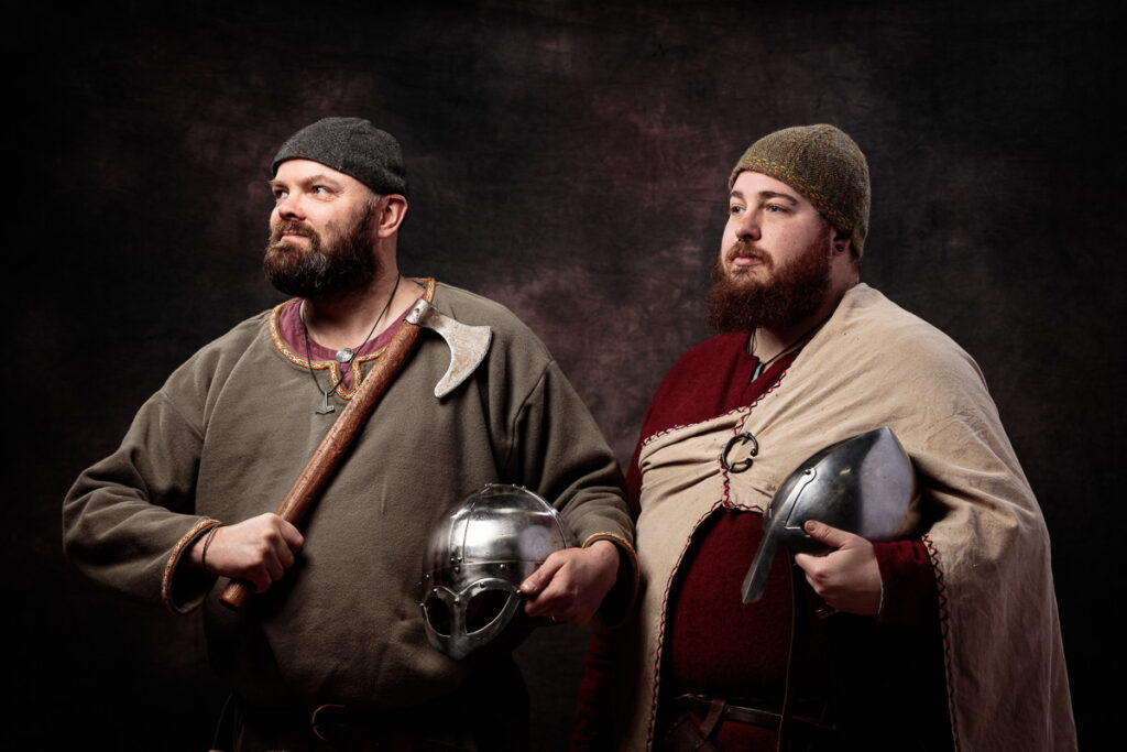 Two bearded men in Viking-era clothing hold axes and helmets against a dark textured background.