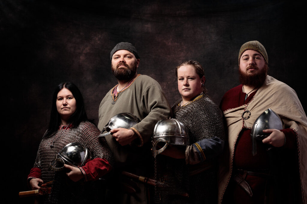 Five people in Viking-era clothing and armour stand against a dark background, each holding metal helmets.