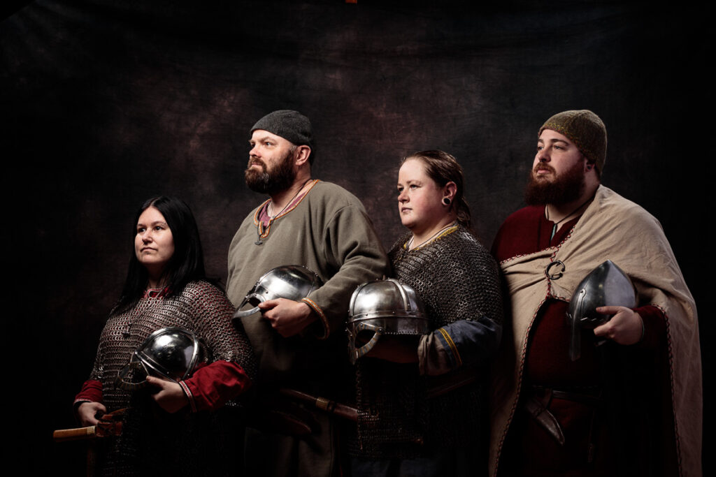 Four people in medieval Viking-style clothing and armour stand against a dark background, each holding a helmet.