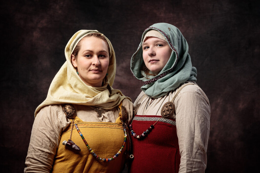 Two women wearing medieval-style clothing and head coverings stand against a dark background, looking toward the camera.
