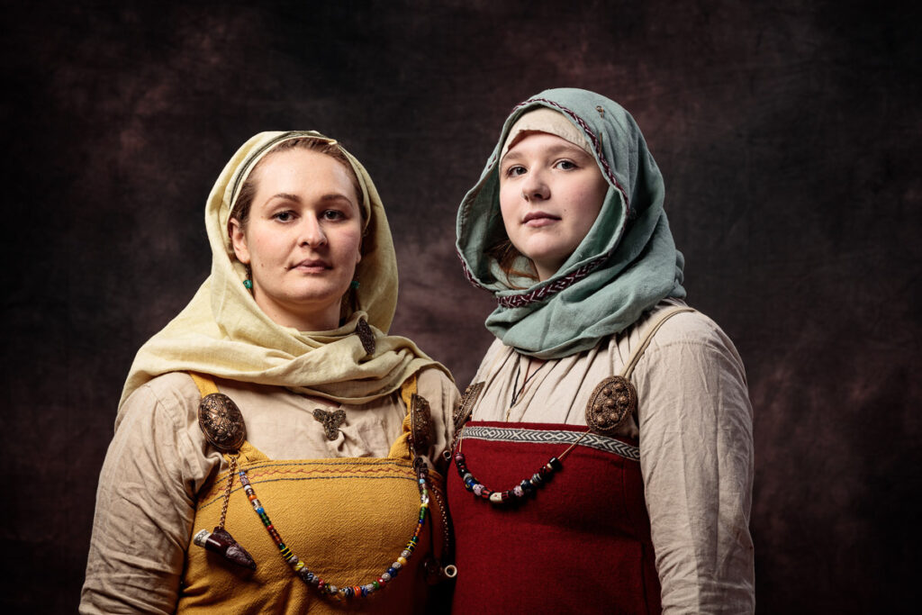 Two women wearing historical Viking-era clothing and head coverings stand against a dark background, looking upward.