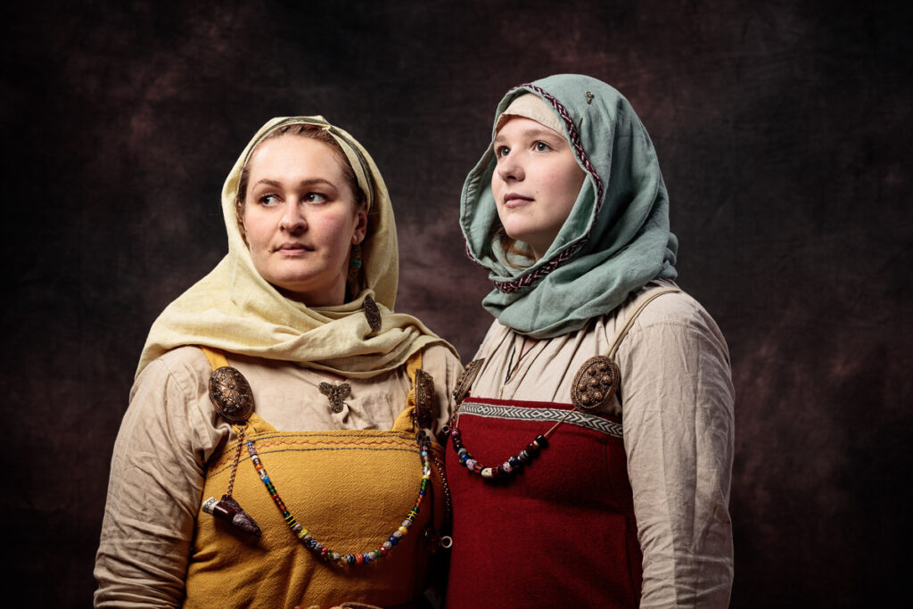 Two women wearing historical Slavic clothing with headscarves and embroidered tunics stand against a dark brown background.