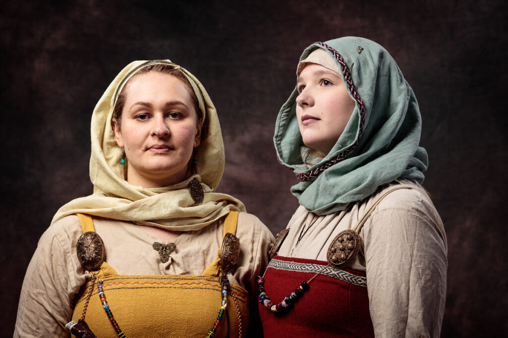 Two women wearing historical Slavic clothing with headscarves and embroidered tunics stand against a dark background.