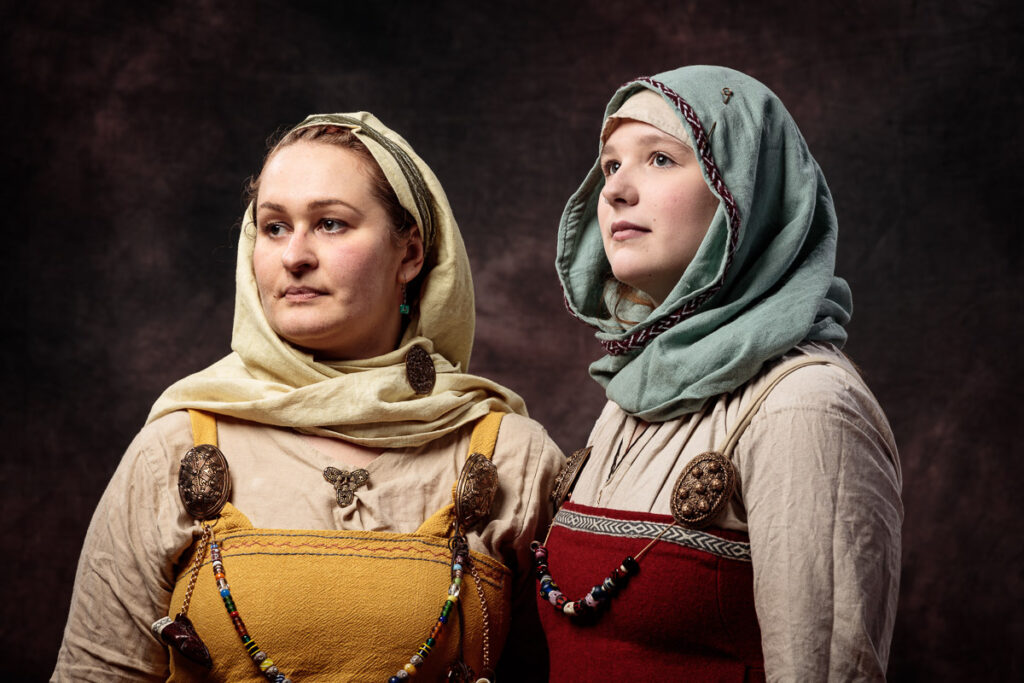 Two women wearing historical Viking-era clothing and head coverings stand against a dark brown background, looking off to the side.