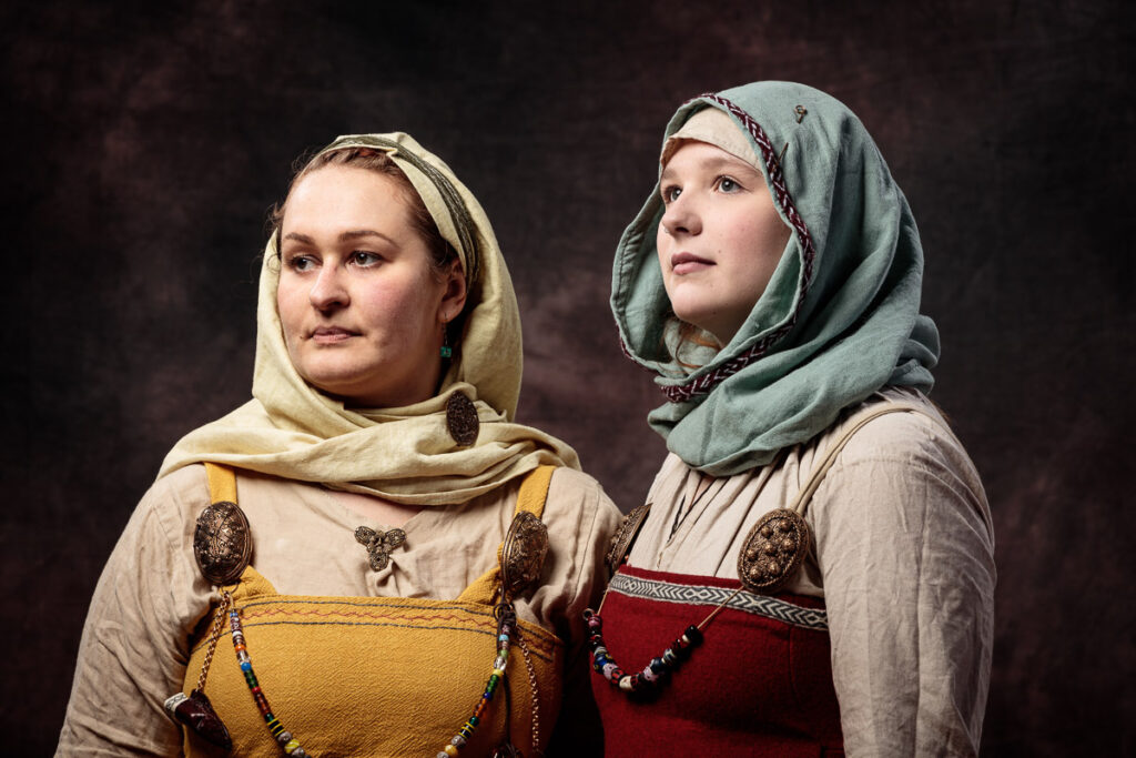 Two women wearing historical Viking or Norse-inspired clothing with head coverings and embroidered tunics against a dark background.