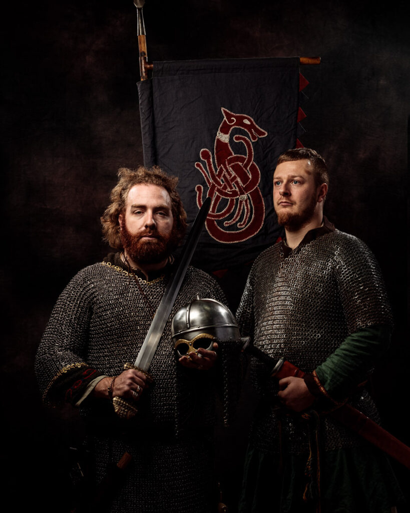 Two bearded men in chain mail armour stand before a dark banner with a red dragon emblem, holding a sword and helmet.
