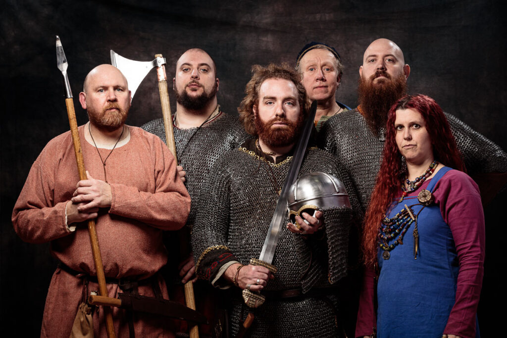 Six people in Viking-era clothing and armour stand against a dark background, holding weapons including axes and swords.