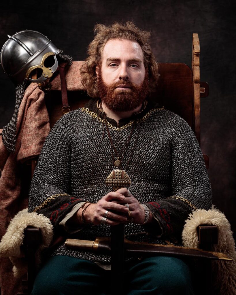 A bearded man in medieval warrior attire sits holding a helmet and axe against a dark background.