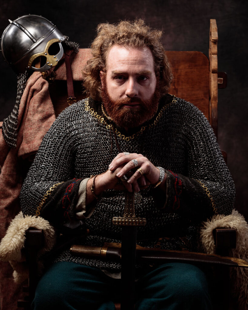 A bearded man in medieval chainmail sits on a wooden chair, holding a helmet. He wears rings and fur-lined garments against a dark background.