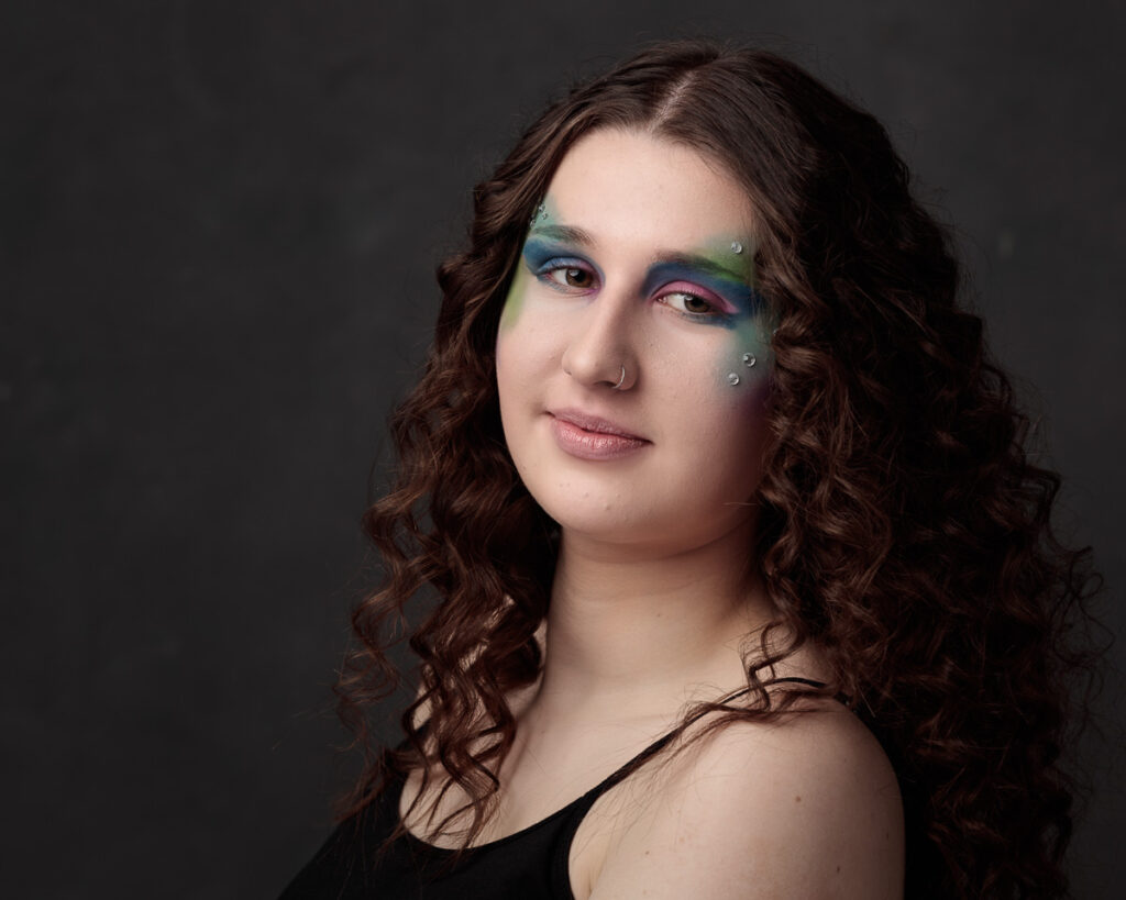 A person with long, wavy brown hair and nose piercing wears colorful eyeshadow in blue, green, and pink against a dark background.